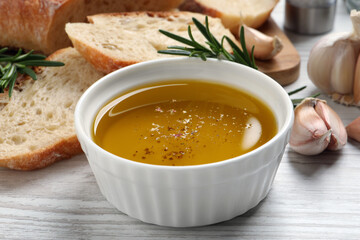 Bowl of fresh oil, bread, rosemary and garlic on white wooden table, closeup