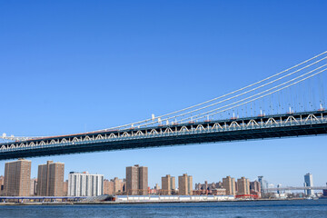Looking up at the span section of Manhattan Bridge on a sunny day with blue sky. High-rise residential buildings behind the suspension bridge deck
