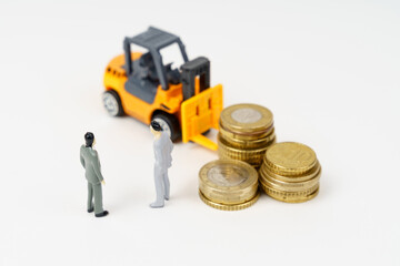 A toy forklift, coins and miniature figures of businessmen stand on a white surface.