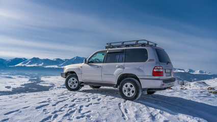 Fototapeta na wymiar The SUV car stands on a snowy plateau. Footprints and shadows in the snow. Ahead and below you can see the river, the mountain range against the blue sky. Altai