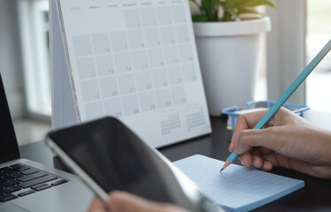 Business women hand with pencil writing note, planning daily appointment and meeting schedule, using mobile phone with year 2022 calendar and laptop on office desk