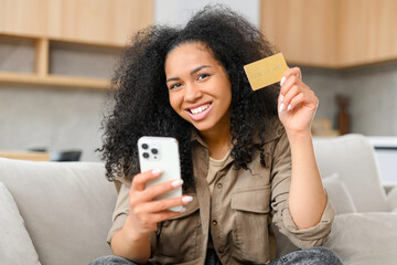 Cheerful young woman using smartphone shopping app for buying online, holding credit card and inputs banking data, sits on sofa in relaxed atmosphere in living room at home