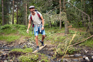 Young hiker in hat with backpack crossing a little creek in the forest. Sport, nature, freedom, holiday concept.