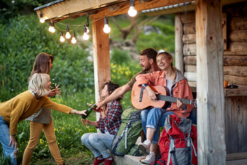 Young cheerful friends and toasting and drinking beer in front of wooden cottage on the terrace. ...