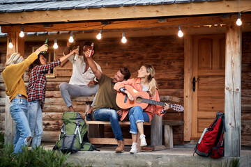 Group of young friends toasting and playing guitar in front of wooden cottage on the terrace.  Summertime garden celebration and fun. Friends, togetherness, fun, joy, celebration concept.