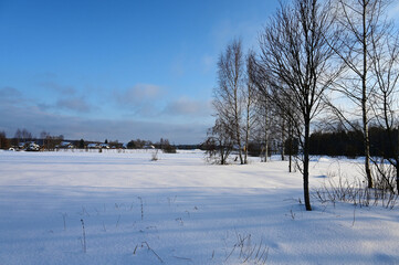 Beautiful winter landscape with village in distance and trees on sunny day. Everything is covered with snow. Sun is shining brightly in blue sky