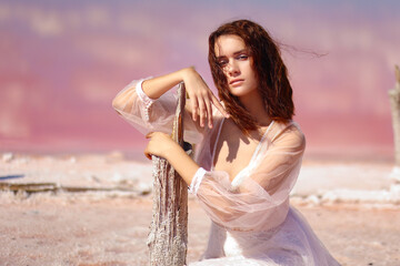 Young woman in white wedding dress sitting in water of pink lake. Romantic portrait of a girl in beautiful landscape.