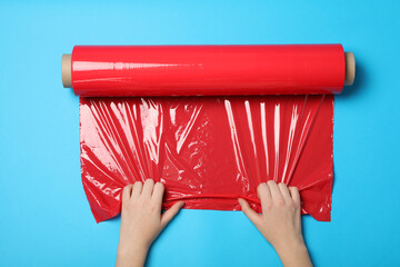 Woman unrolling red plastic stretch wrap on light blue background, top view - obrazy, fototapety, plakaty