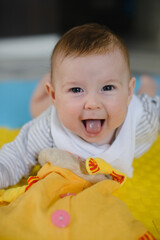 The baby is lying on his stomach on the mat and playing with toys. He looks at the camera and smiles.