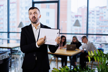 An adult businessman with a folder in his hands stands in the office against the background of his employees.