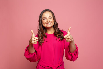 Young happy woman smiling showing thumbs up because something good has happened over pink background