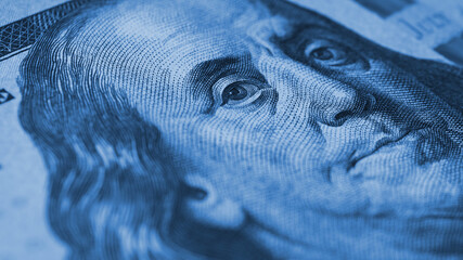 American paper money background. A $100 bill with focus on Benjamin Franklin eye. US banknote close up. Dark blue tinted wallpaper about economy and the USA dollar. Macro