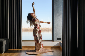 Attractive young woman dressed in a dressing gown dances in front of a huge window in the room. The girl raises her arms above her head.