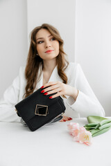 High fashion portrait of a young blonde model in a classic white suit. Vogue style. The girl sits at a table against a white background and holds a small leather handbag. 