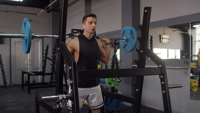 Man of athletic build in sportswear and headphones trains at gym doing squats with barbell, front view. Man of athletic build trains in gym with barbell