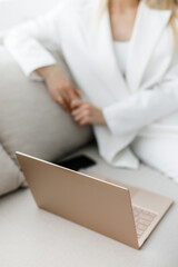 A businesswoman in a white suit works on her gold laptop. a blonde girl sits on the sofa and takes notes on her laptop. 