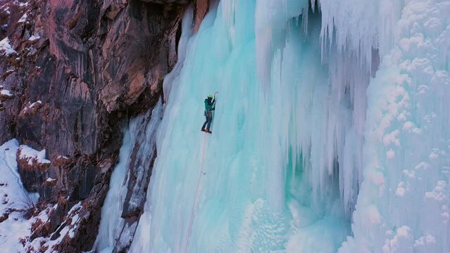 Mountaineer Man is leading on Ice. Ice Climbing on Frozen Waterfall, Aerial View. Barskoon Valley, Kyrgyzstan. Drone Flies Upwards, Tilt Down. Crane Shot