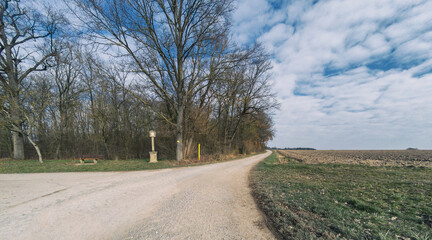 Ein Bildstock und eine Bank an einem Waldstück neben Feldern und einem Feldweg