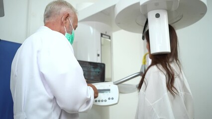 Selective focus side view of a Caucasian senior dentist in a face mask, pressing buttons on a 3D panoramic teeth x-ray machine while helping a teenage patient received a teeth x-ray in dental clinic. - Powered by Adobe