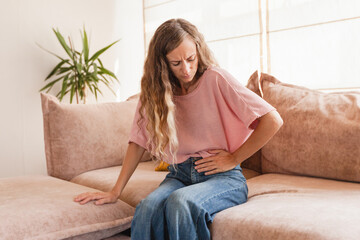 Young woman suffering from strong abdominal pain while sitting on sofa at home