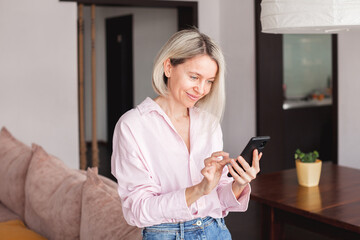 Attractive middle-aged 40s woman looking at her smart phone and smiling while standing at home