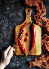 Flatlay with red hot chili peppers and its slices on a wooden cutting board with a hand which holds a knife, fabric on a black concrete background