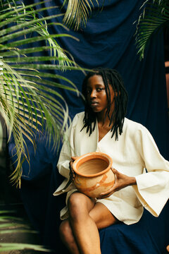 Portrait of a young african indigo eco fashion designer holding pottery in front of a blue background