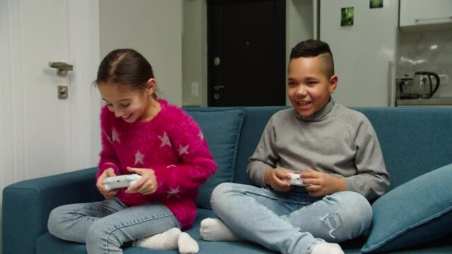 Multiethnic preteen young friends competing by playing video game while sitting on sofa indoors. Cheerful white girl and cute black boy spending leisure time together, having fun at home. Moving shot