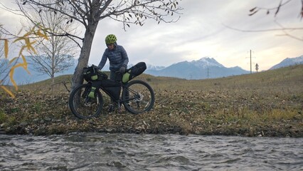 Fototapeta na wymiar The woman travel on mixed terrain cycle touring with bikepacking. The traveler journey with bicycle bags. Sportswear in green black colors. The trip in magical autumn forest, river stream.