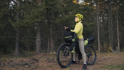 Naklejka na ściany i meble The woman travel on mixed terrain cycle touring with bike bikepacking outdoor. The traveler journey with bicycle bags. Sportswear in green black colors. Magic forest park. Make a selfie smartphone.