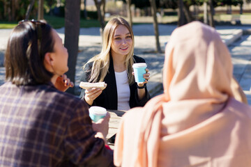 Three diverse girls having coffee break and chatting in city park