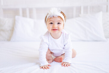 cute baby girl sitting on the bed in white clothes and with a bow on her head, funny little baby on a cotton bed at home and smiling, baby products concept