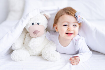 funny joyful baby girl looks out from under the blanket on the bed in white clothes with a teddy bear in her hands and smiles, a small child on a cotton bed at home woke up after sleeping