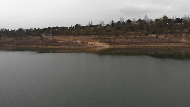 Aerial footage approaching the shore of a reservoir at very low altitude. Cloudy day with rain clouds.