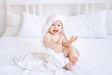 a joyful baby girl of six months on a bed with a white towel on her head after bathing or washing,...