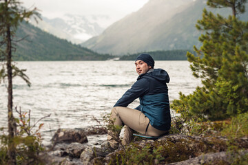 A male hiker and adventurer sits on the shore of a picturesque mountain lake and admires the landscape.