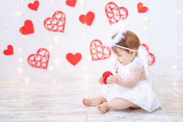 Valentine's day concept, a joyful baby girl of six months in an angel costume with wings and a red heart in her hands smiles