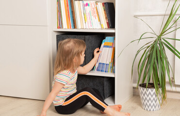 Little girl chooses a book in a bookcase at home. The child looks through the books in the library,...