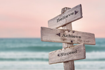 believe achieve suceed text quote written on wooden signpost by the sea. Positive pink turqoise pastel theme.