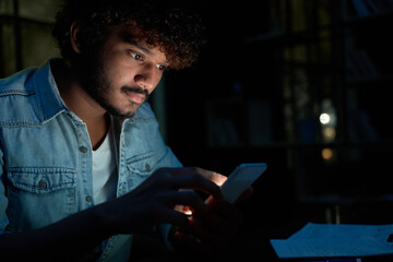 Young indian man using mobile apps technology on cell phone late at night searching information in...