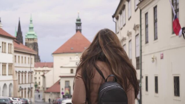 Young woman walking Prague with backpack and camera, turning around, middle shot guide view