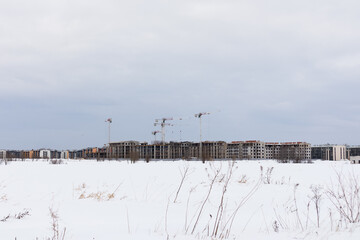 Tower cranes on construction of buildings on snowy field under cloudy sky, view from afar