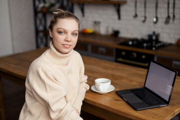 Happy young woman is in a cozy home atmosphere, sits at the kitchen table and holds a cup of coffee in her hands