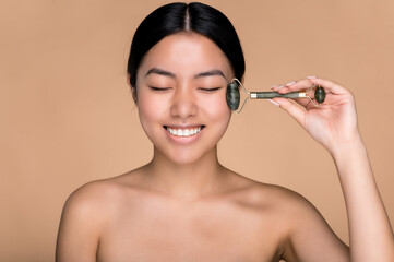 Fototapeta premium Asian beautiful girl holding a jade massage roller for facial skin in her hand, makes anti-aging measures, wrinkle prevention, standing on an isolated beige background, smiling with eyes closed