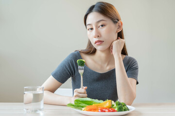 Obraz na płótnie Canvas Diet in bored, unhappy beautiful asian young woman, girl on dieting, holding fork at broccoli in salad plate, dislike or tired with eat fresh vegetables. Nutrition of clean, healthy food good taste.