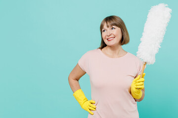 Elderly smiling housewife woman 50s in pink t-shirt gloves doing housework hold white duster brush look aside isolated on plain pastel light blue background. Housekeeping cleaning tidying up concept.