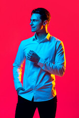 Portrait of young handsome man in white shirt smiling, looking away, posing isolated over red sttudio background in blue neon light