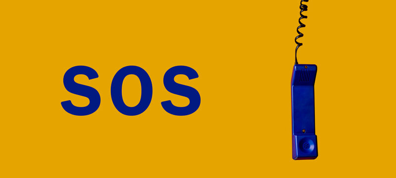 Old blue telephone hanging from above on one side, on a yellow background with the word SOS in blue color on the other side, panoramic photography.