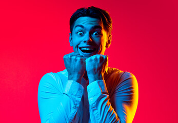 Portrait of young emotive man, student in white shirt showing emotions of excitement isolated over red studio background in blue neon