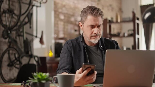 Bearded man working online with laptop computer and phone at home sitting at desk. Home office, browsing internet. Portrait of mature age, middle age, mid adult man in 50s.